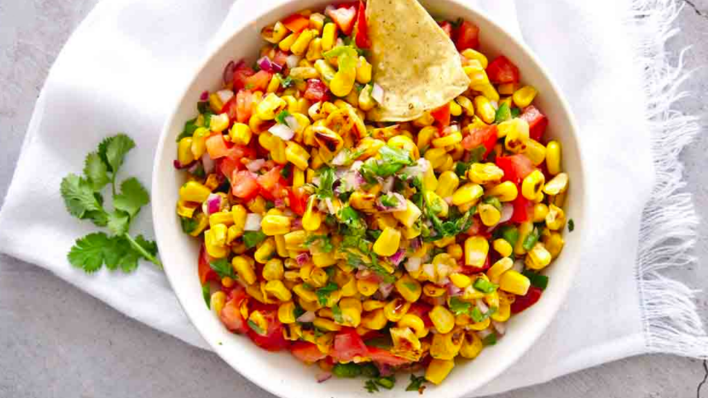 An overhead view of a white bowl filled with corn salsa. It sits on a white cloth with a sprig of fresh cilantro to the left side.