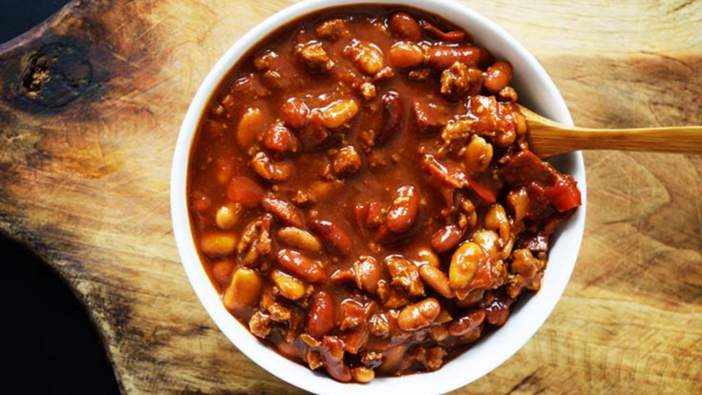 An overhead view of a white bowl full of cowboy beans. A wood spoon rests in the side of the bowl.