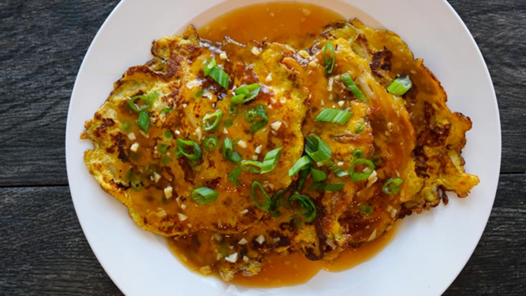 An overhead view of a white plate holding 4 egg foo young pancakes which are drenched in sauce and garnished with fresh, sliced, green onions.