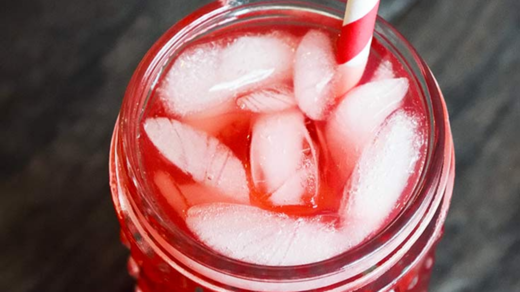 An up close view of a glass of hibiscus lemonade. A red and white stripped straw sits between ice cubes in the drink.
