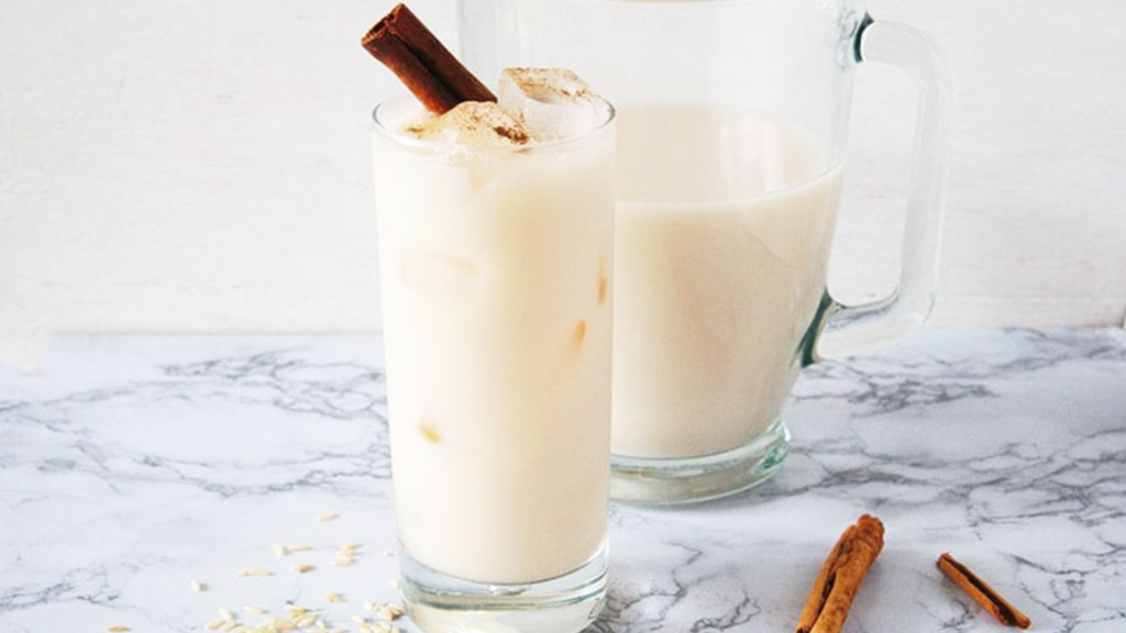 A glass sits on a marble surface filled with homemade horchata and is garnished with a cinnamon stick. A pitcher of horchata sits behind the glass.