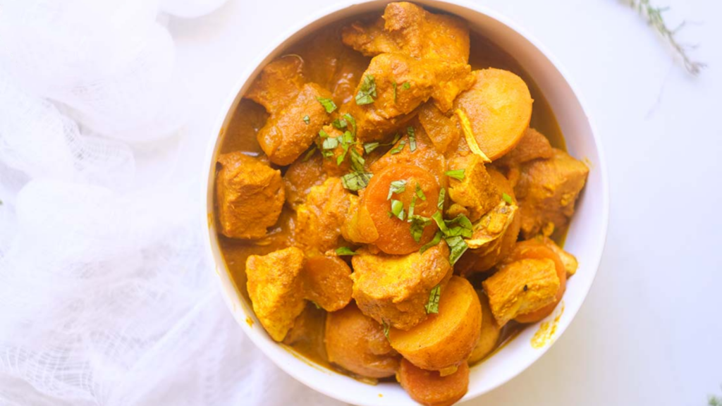 A white bowl holds Jamaican Curry Chicken with large chunks of chicken, potatoes and carrots.