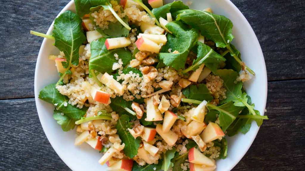 An overhead view of a white bowl holding kale quinoa salad with apple pieces.