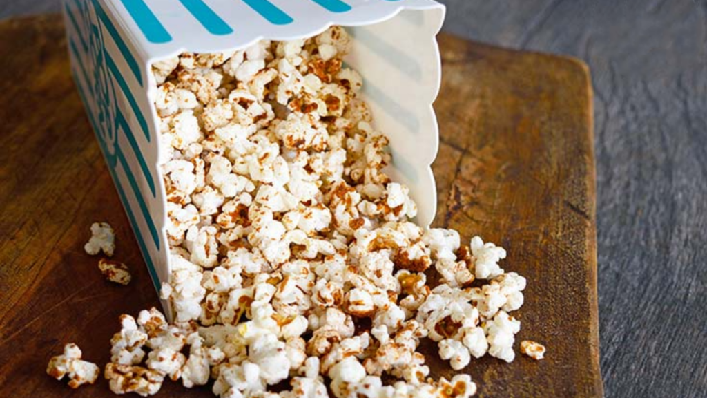 A tipped over popcorn container spills homemade kettle corn onto a wood cutting board.