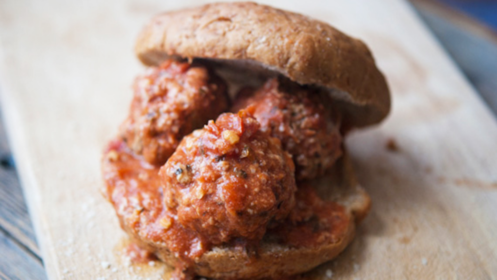 A single meatball sandwich on a whole grain bun sits on a wood cutting board.