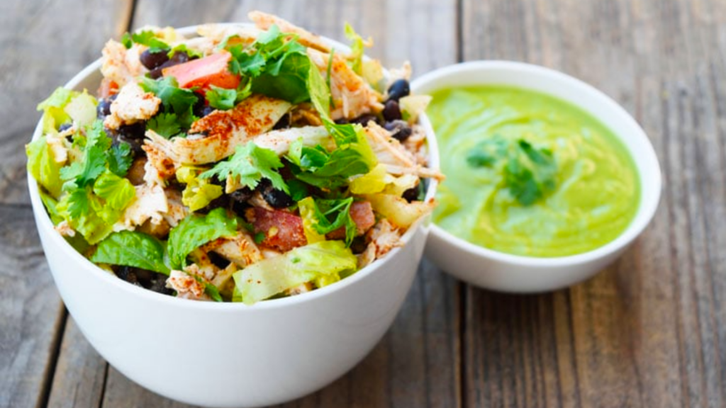A white bowl sits filled with Mexican Chicken Salad on a wood table. A smaller white bowl of avocado dressing sits next to it.