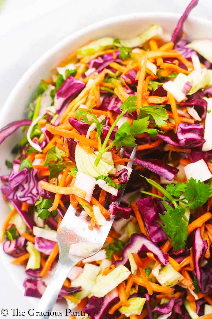 A closeup of Mexican Coleslaw in a white bowl. A fork lifts a bit of it up out of the bowl.