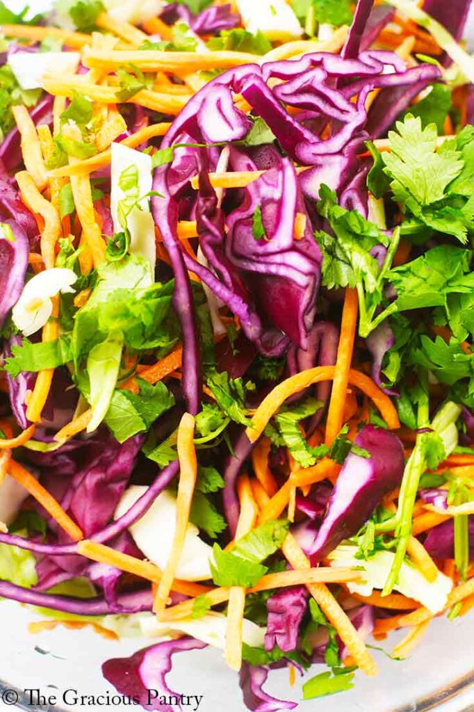 A closeup of purple cabbage, shredded carrots and greens in a glass bowl.