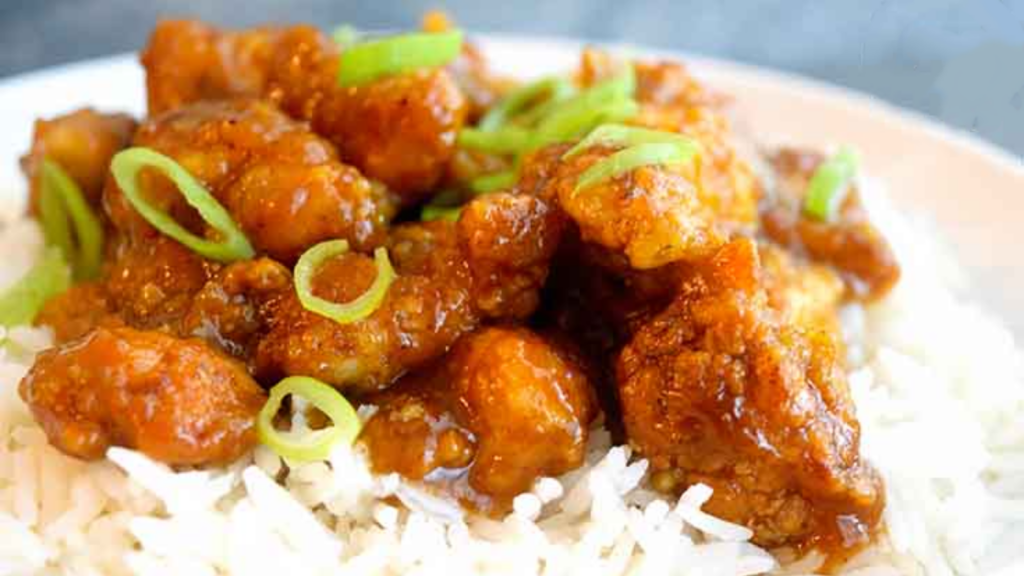 A closeup of orange chicken garnished with sliced green onions and laying on a bed of rice.