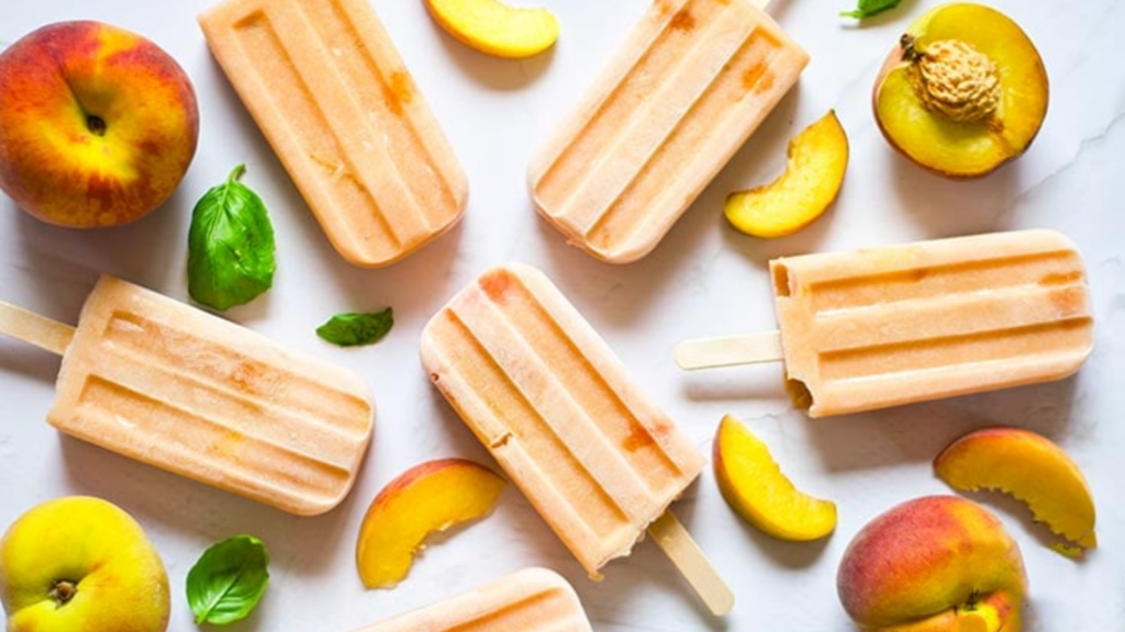 Peach popsicles laying on a white surface. Pieces of peaches lay around them.