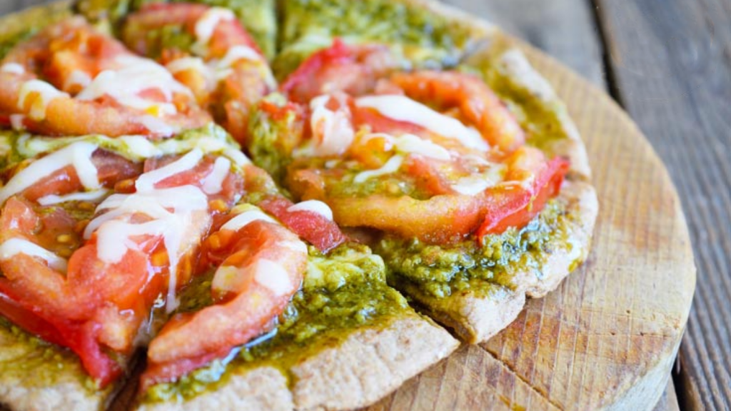 A pita sits on a small round cutting board covered in pesto sauce, fresh tomato slices and melted cheese.