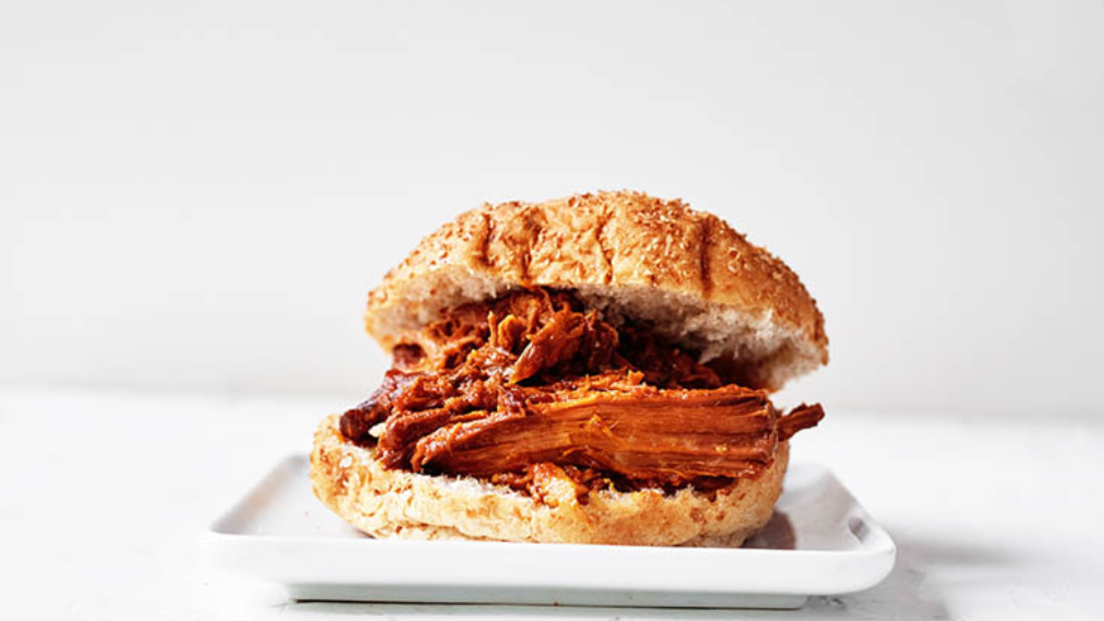 A single pulled pork sandwich sits on a square, white plate on a white background.