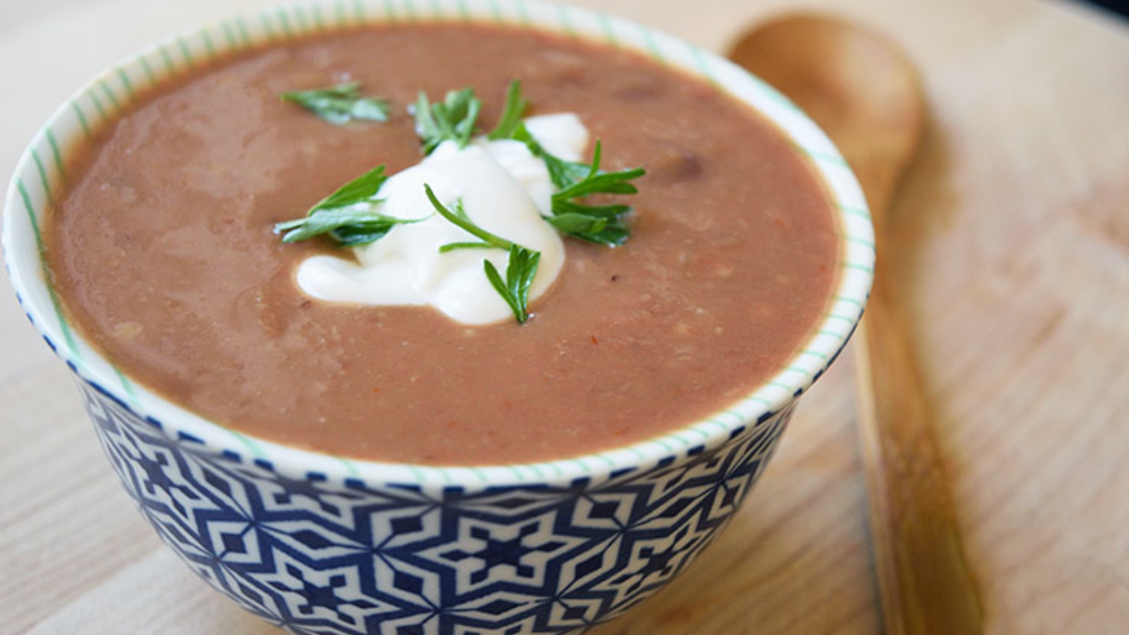 A small bowl of Refried Bean Soup on a wood table. A wood spoon lays next to the bowl.