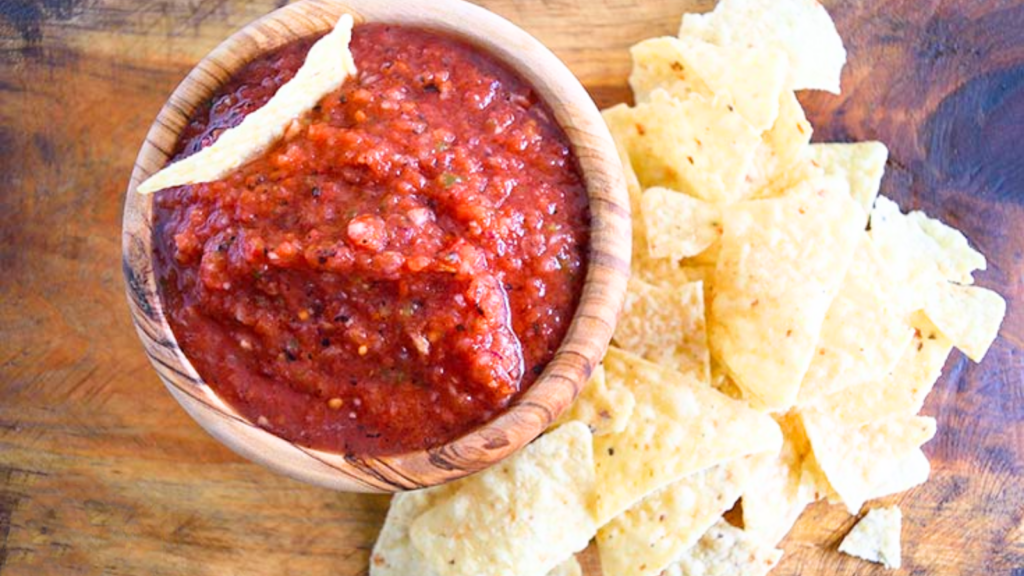 An overhead view of a wood bowl filled with restaurant salsa. Chips lay on a wooden surface to the right of the bowl.