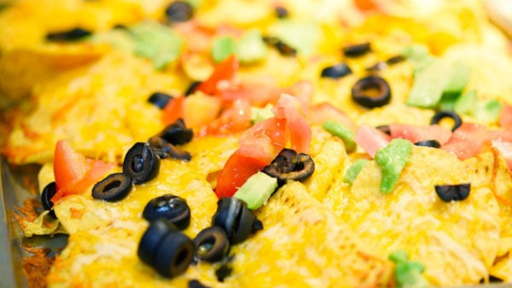 Sheet pan nachos on a sheet pan with melted cheese over the top, and garnished with olives, avocado pieces and chopped tomatoes.