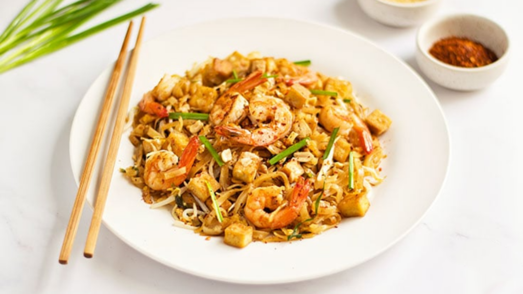 A white plate holds a serving of shrimp pad thai. Chopsticks rest on the side of the plate.
