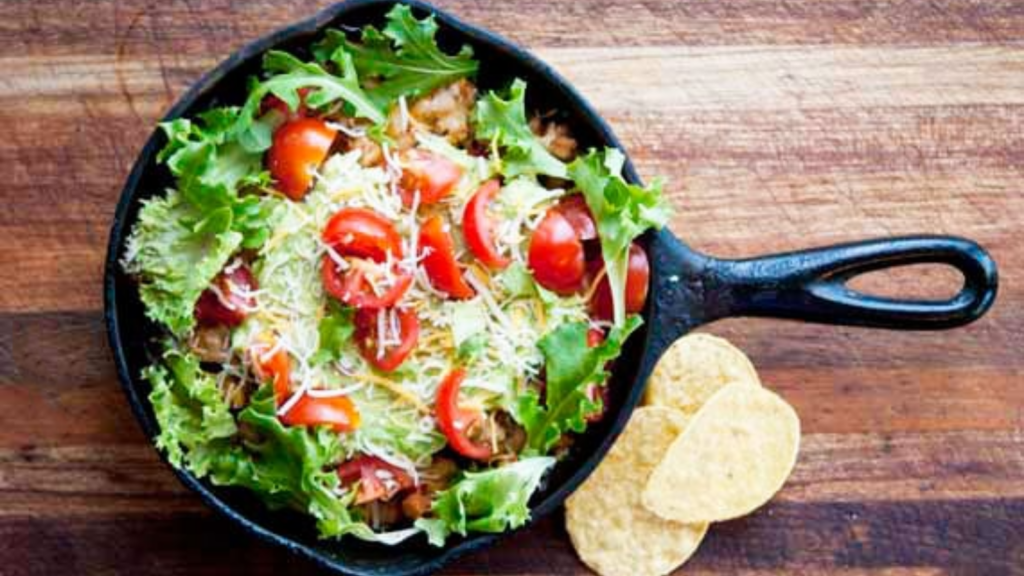 An overhead view of a cast iron skillet holding a skillet taco salad topped with shredded cheese and halved grape tomatoes.