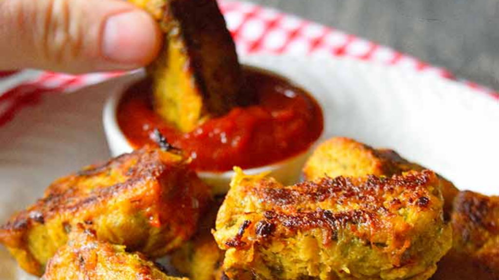 A hand dips a spaghetti squash tot into a small dish of ketchup behind a plate holding a big pile of them.