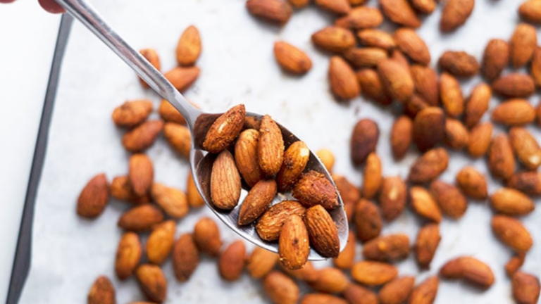 A spoon lifts up a spoonful of spicy roasted almond from a baking sheet full of them.