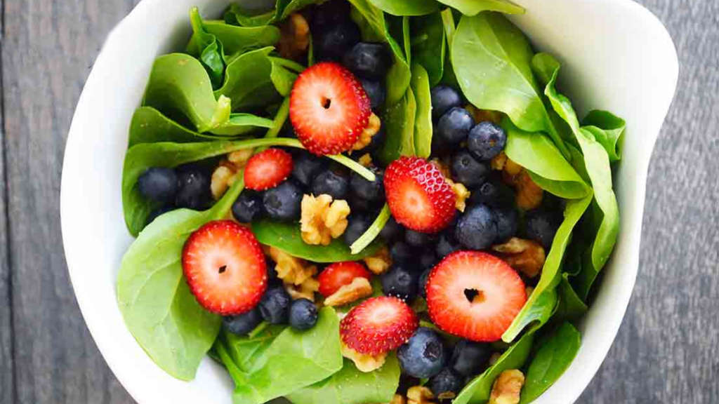 And overhead view of a white bowl filled with spinach salad. The spinach is topped with fresh strawberries, blueberries and pieces of walnuts.