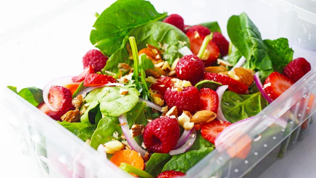 A plastic container holds a fresh looking salad filled with greens, raspberries, strawberries and more.
