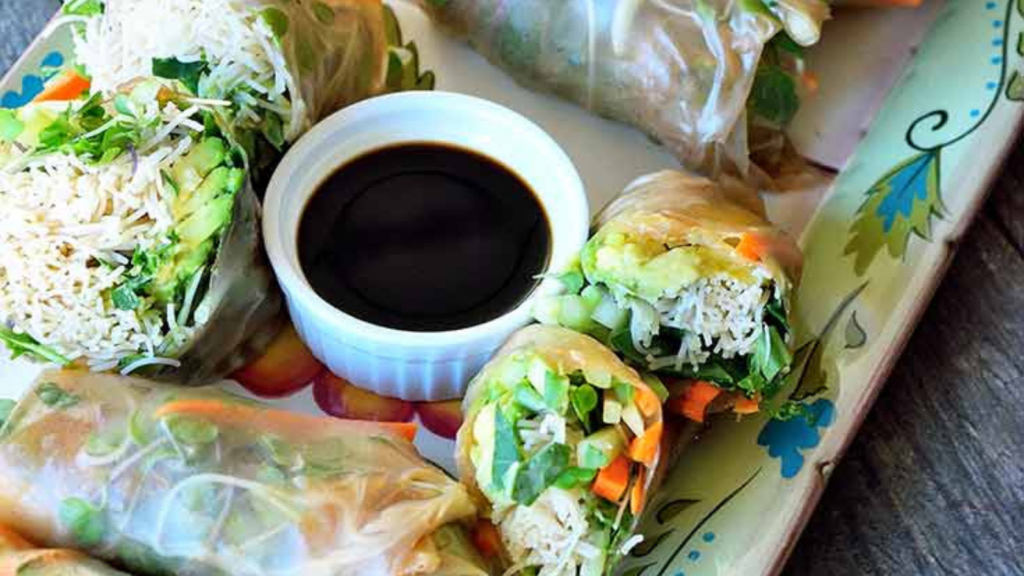 A decorative platter is filled with fresh spring rolls surrounding a small white bowl of soy sauce.