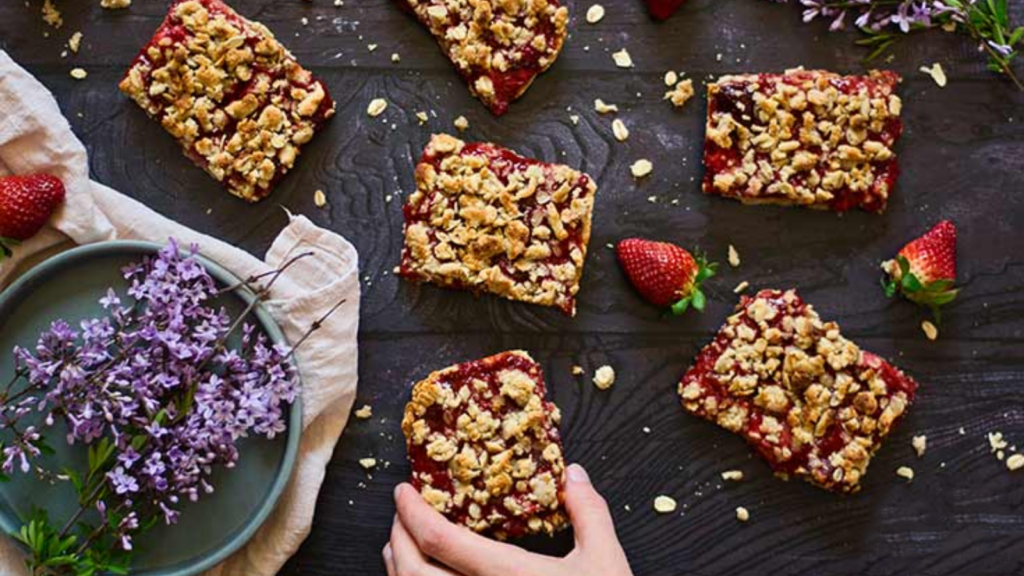 A hand reaches for a strawberry crumb bar from a group of them on a table.