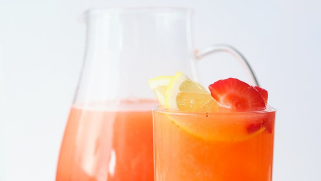 A closeup of a glass filled with strawberry lemonade and a pitcher standing right behind it.