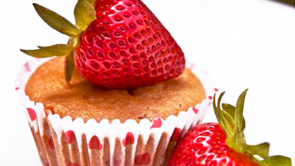 A strawberry muffins with a fresh strawberry laying on top of it.