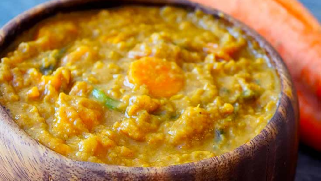 A wood bowl sits filled with sweet potato curry. Two large raw carrots lay next to the bowl.