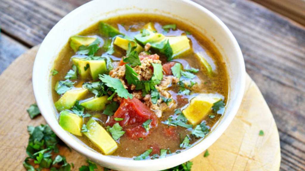 A white bowl filled with low carb taco soup, garnished with avocado chunks and fresh cilantro.