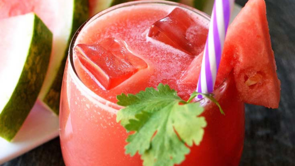 A closeup view of a glass filled with watermelon lemonade and garnished with a cilantro leaf, watermelon wedge and purple-striped straw.