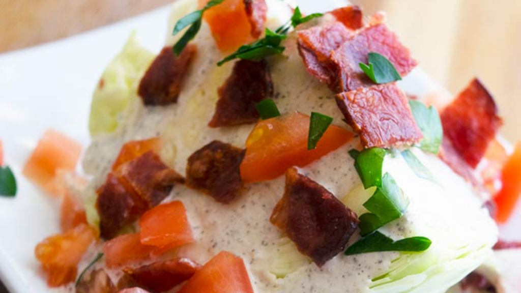 A salad wedge sits on a white plate smothered in bacon bits, chopped tomatoes and ranch dressing.