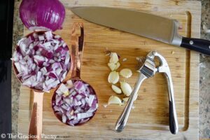 Chopped red onion in two measuring cups on a wood cutting board next to some garlic cloves, a garlic press and a knife.