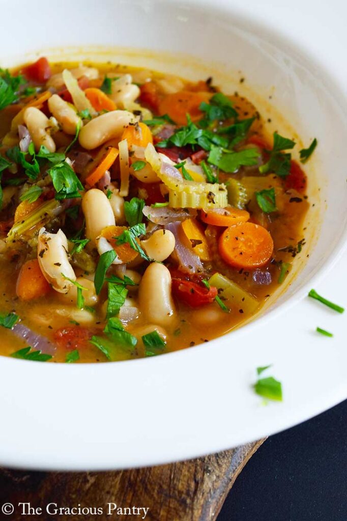 A side view of a wide-rimmed, white bowl holding a serving of white bean soup that is garnished with fresh, chopped parsley.