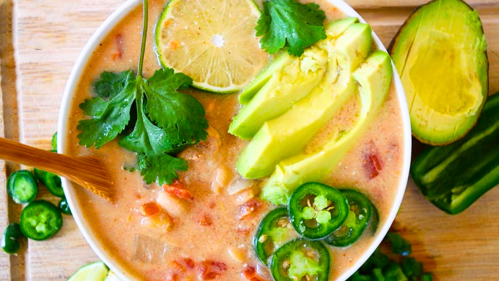 An overhead view of a white bowl filled with white chicken chili and garnished with fresh cilantro, avocado slices and jalapeno slices.