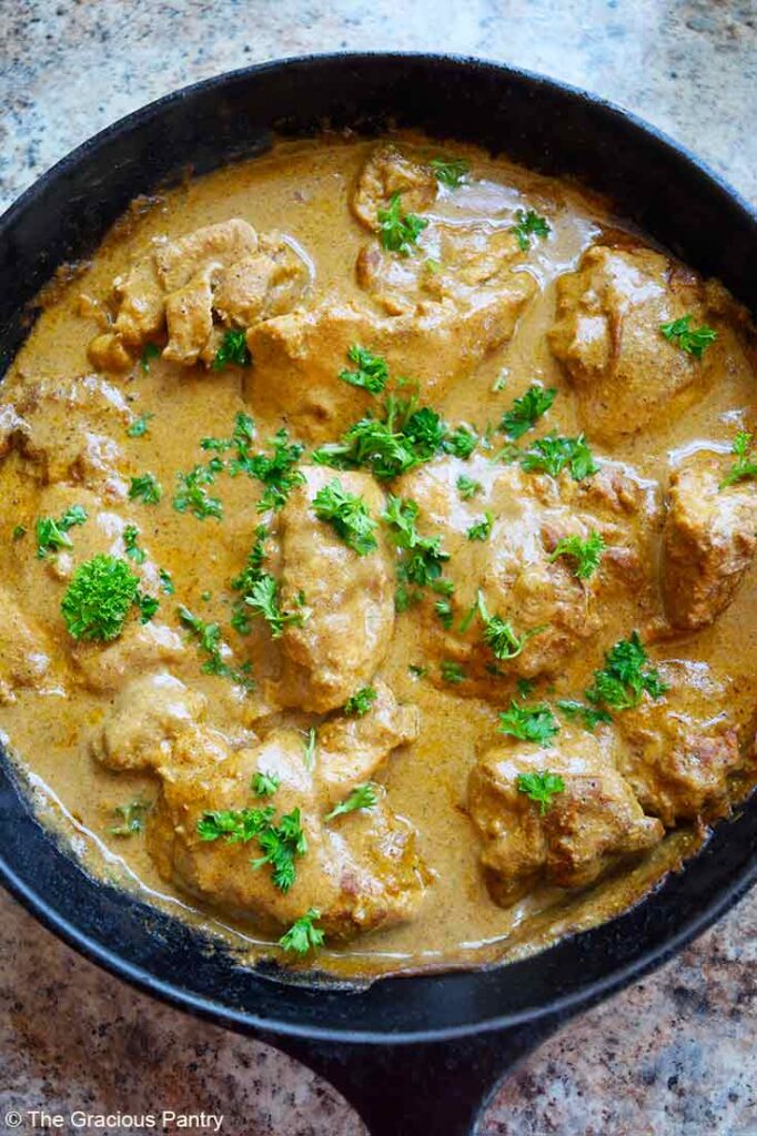 An overhead view of a cast iron skillet show a pan full of Yogurt Chicken, baked and garnished with fresh, chopped parsley.