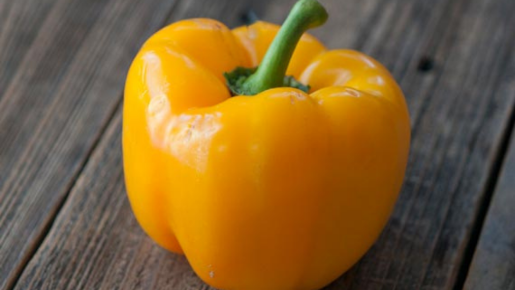 A single yellow bell pepper sitting on a wooden surface.