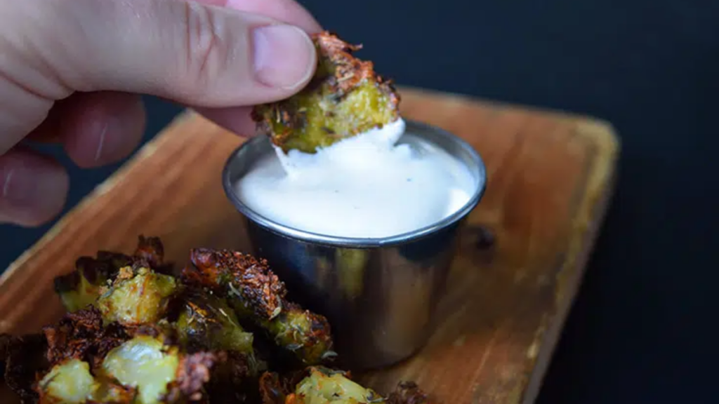 A hand dipping a smashed brussels sprout into a small bowl of ranch dip.