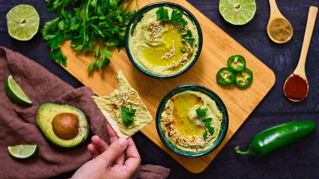 A hand holds a cracker with avocado hummus on it. Two bowls of avocado hummus sit on a table below.