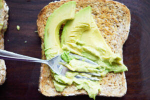 A fork smashing avocado slices into a slice of toast.