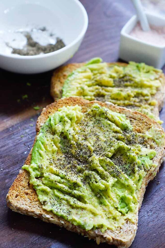 Two slices of avocado toast on a dark wood surface with two white dishes behind them holding salt and pepper.