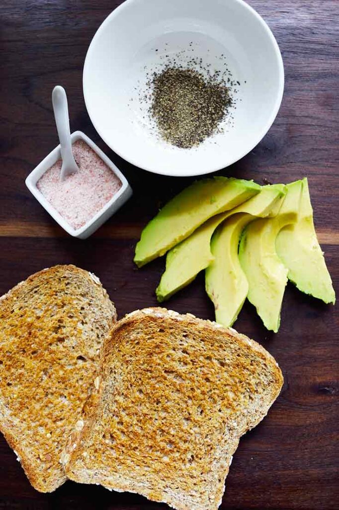 An overhead view of all the avocado toast recipe ingredients gathered on a dark wood surface.