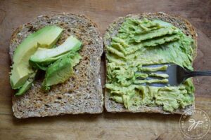 Two slices of toast. One has avocado slices on it and the other is having avocado mashed on it with a fork.