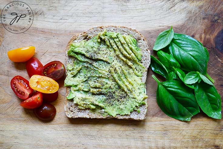 A prepared avocado toast with tomato halves on the left and basil leaves on the right.