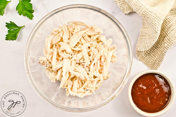 Shredded chicken in a glass bowl with a small white bowl to the right containing barbecue sauce.