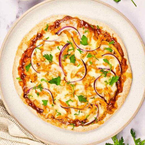 A BBQ Chicken Pizza lays on a plate with a small bowl of bbq sauce off to the top left and parsley sprinkled all around the plate.