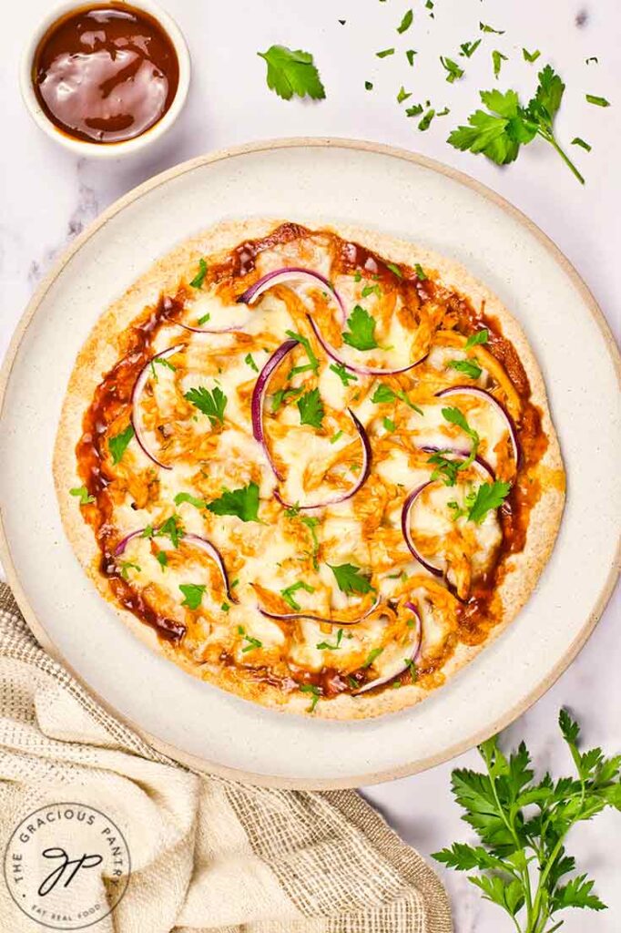 A BBQ Chicken Pizza lays on a plate with a small bowl of bbq sauce off to the top left and parsley sprinkled all around the plate.