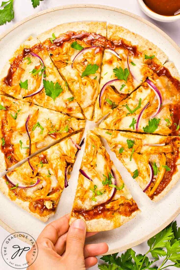 A hand reaches for a slice of BBQ Chicken Pizza off of a plate.