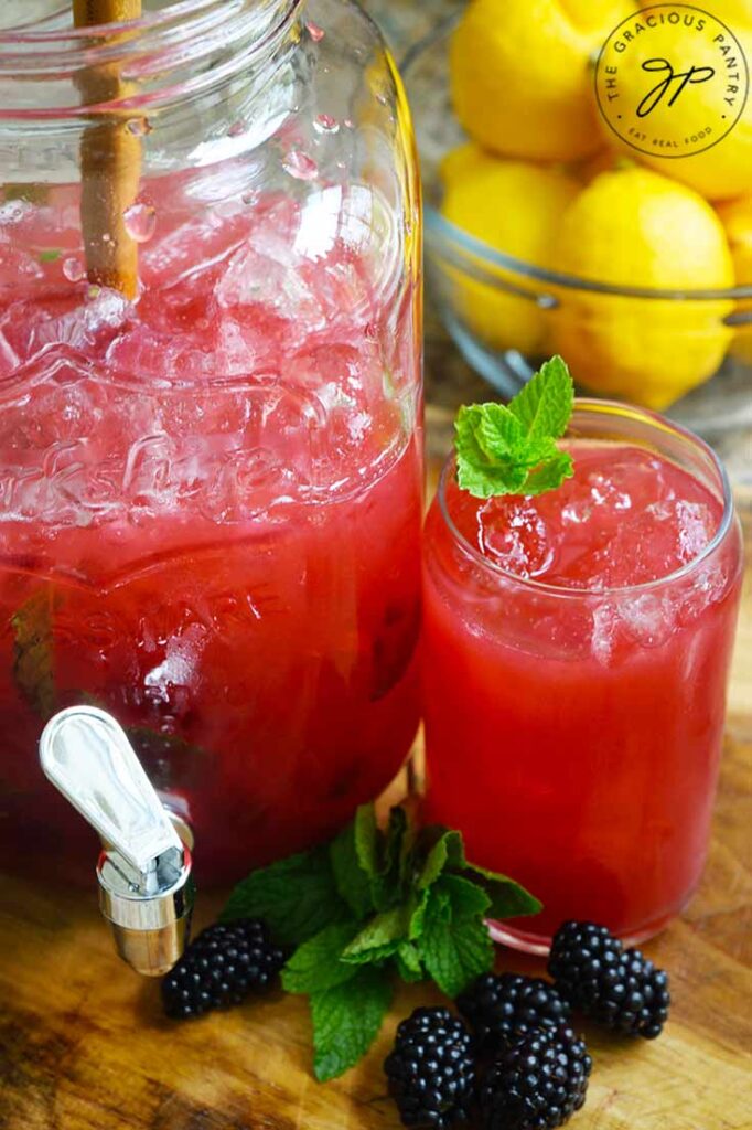 A glass of Blackberry Lemonade sits next to a large beverage jug filled halfway with the same.
